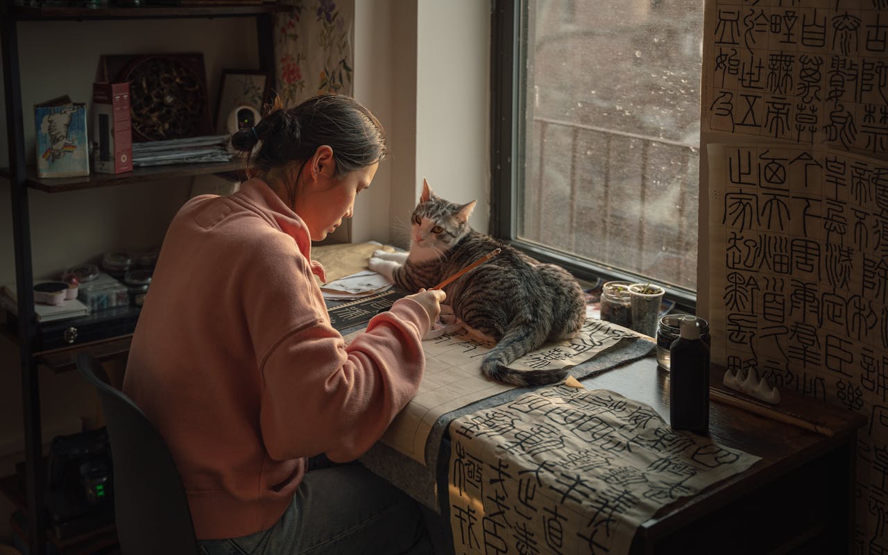 services-02 A woman focuses on Chinese calligraphy beside a cat in a cozy New York apartment.