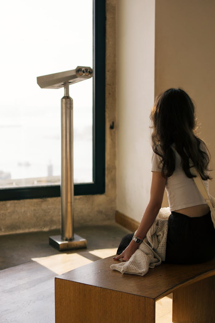 Woman sitting near a window, contemplating view with a telescope beside her, indoors.